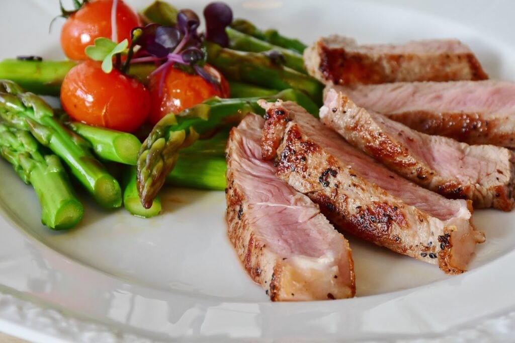 Veggie vs. Meat: Fight or Feast? 
A white plate with a grilled beef steak in the center. To the left of the steak are asparagus spears, and to the right is a sliced tomato.